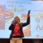 Vivianne Verschae ofreciendo una charla con micrófono en mano frente a una presentación proyectada sobre creencias y roles sociales.