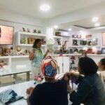 Vivianne Verschae impartiendo un taller presencial a un grupo de mujeres, rodeada de artesanías y pantallas con su imagen promocional.